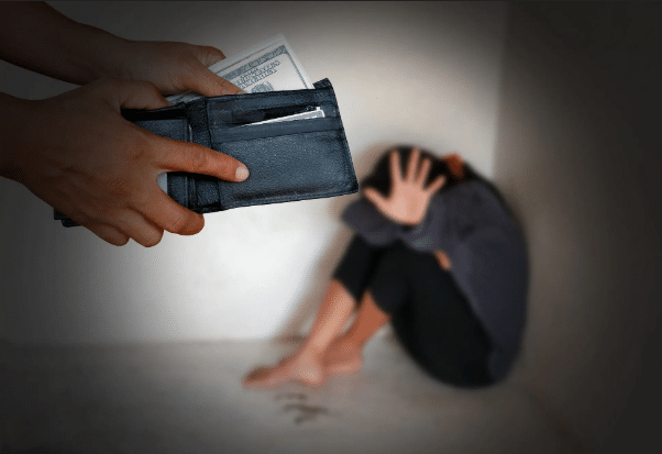 A person's hand holds an open, dark wallet with US dollars visible inside toward a distressed young woman who is sitting barefoot, huddled in a corner with her face obscured, holding one hand up in a defensive or fearful gesture. The lighting is dim, suggesting a dark, possibly confined space, and there is a stark contrast between the foreground action and the background figure. The image suggests themes of coercion, exploitation, or human trafficking, particularly in relation to money.